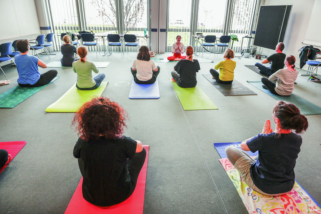 Eine Yogagruppe sitzt auf vielen bunten Yogamatten in einem großen Raum.