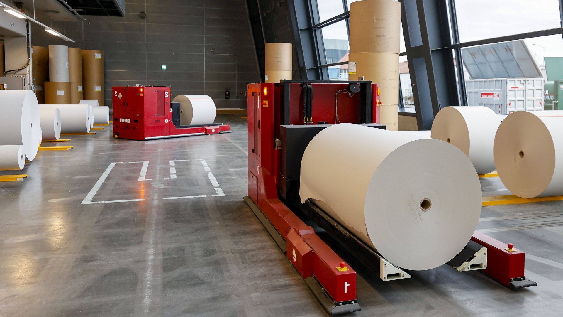 Zwei rote Maschinen transportieren große Rollen an weißem Papier im Druckzentrum.